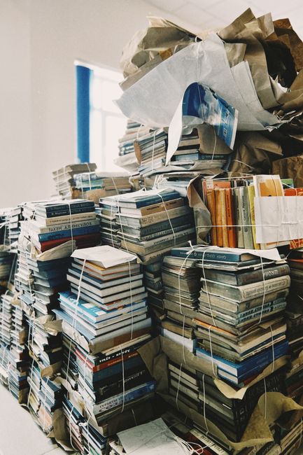 A large collection of tightly bound stacks of books and magazines, secured with white plastic strapping, are positioned in an indoor setting, likely a storage or office space. The books vary in size, 