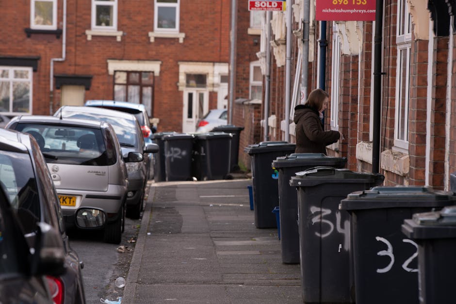 A worker in a bright orange and yellow high-visibility jacket is operating a large red rubbish collection truck on a roadside, with the truck's rear loading mechanism open and partially extended, reve